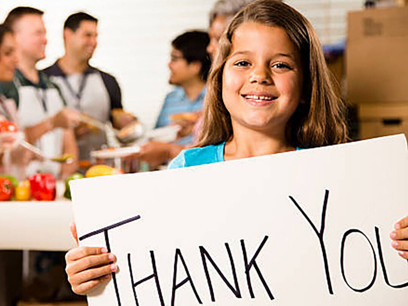 Multi-ethnic and mixed-age group of volunteers serve food to needy people at a community "soup kitchen" or food pantry location. A little girl, who received services, is in the foreground holding a "Thank You" sign. The volunteers wear aprons, lanyards, and gloves. Vegetable, fruit, and soup food items are on the table with boxes all around the group.  Service, kindness, charity, community outreach